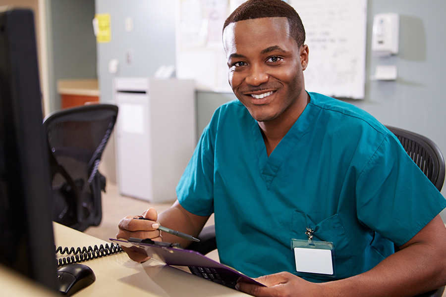 Smiling healthcare worker in teal scrubs sitting at a desk, writing on a clipboard beside a computer monitor.